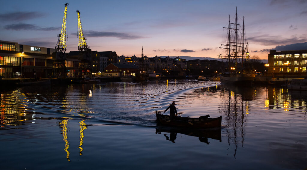 A man and a dog sweep across Bristol harbour in a boat at night